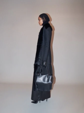 A slender adult female poses indoors in a long black coat with fur trim, a black headdress, black gloves, and carrying a small black handbag.