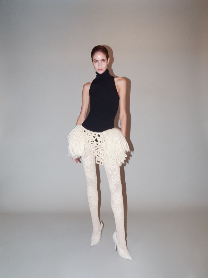 Full-length shot of a woman in a black turtleneck, textural miniskirt, and patterned tights in a studio setting on a neutral backdrop.