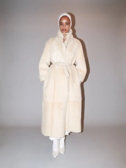 Adult woman wearing a long, light-beige belted coat and matching head covering poses in a photo studio with a solid grey backdrop.