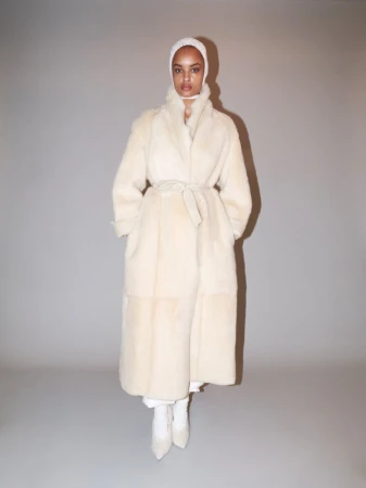 Adult woman wearing a long, light-beige belted coat and matching head covering poses in a photo studio with a solid grey backdrop.