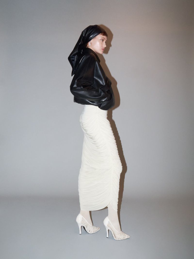 Woman posing in black leather jacket, headscarf, white ruched skirt, and embellished heels against a neutral grey background.