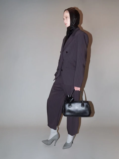 Woman in pinstripe suit, black headscarf, and grey heels holds a black purse against a grey backdrop in a studio shot.