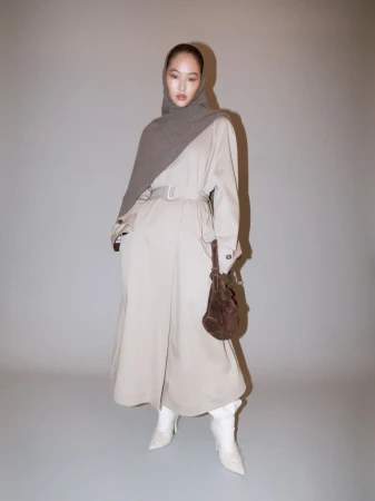 Fashionable woman poses in a tan trench coat, gray headscarf, and white boots, standing against a neutral background in a studio setting.