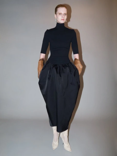Full shot of a fashion model in a black turtleneck, skirt, fur sleeves, and heels stands against a neutral studio backdrop.