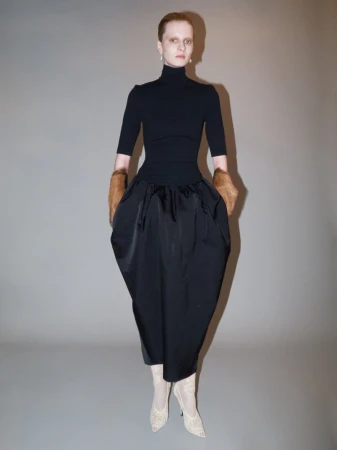 Full shot of a fashion model in a black turtleneck, skirt, fur sleeves, and heels stands against a neutral studio backdrop.