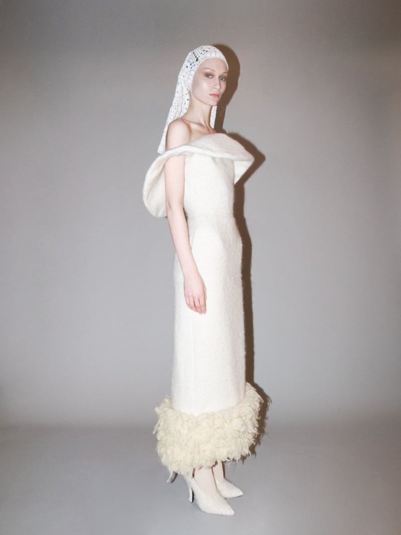 Elegant woman in a white off-the-shoulder dress and lace head covering, posing in a studio with soft, neutral lighting.