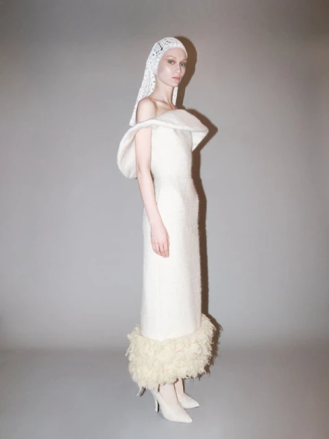 Elegant woman in a white off-the-shoulder dress and lace head covering, posing in a studio with soft, neutral lighting.