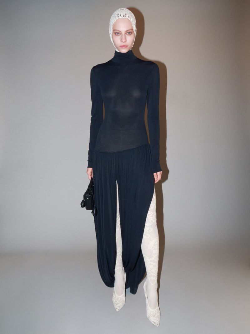 A woman in a long-sleeved black turtleneck, wide-leg pants, lace head covering and lace boots, holding a small black bag, stands indoors.