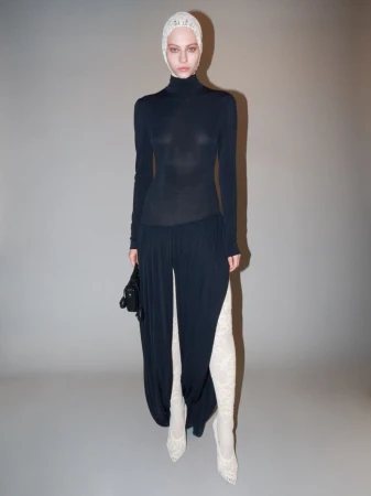 A woman in a long-sleeved black turtleneck, wide-leg pants, lace head covering and lace boots, holding a small black bag, stands indoors.
