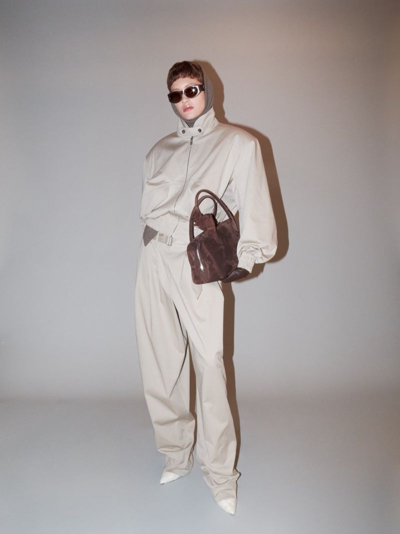 Adult woman wearing a beige jumpsuit, sunglasses, and white heels holds a brown purse in a studio setting with a grey background.