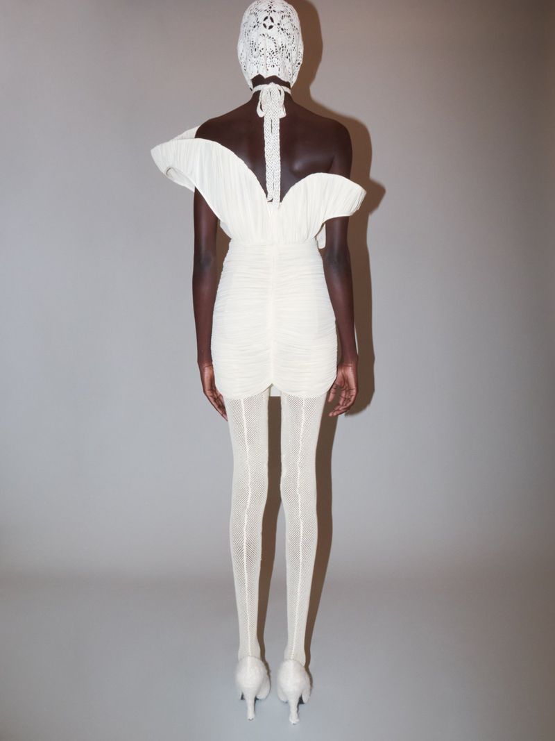 Dark-skinned fashion model in a short ruched dress, textured hose, and high heels posing in a studio against a light grey background.