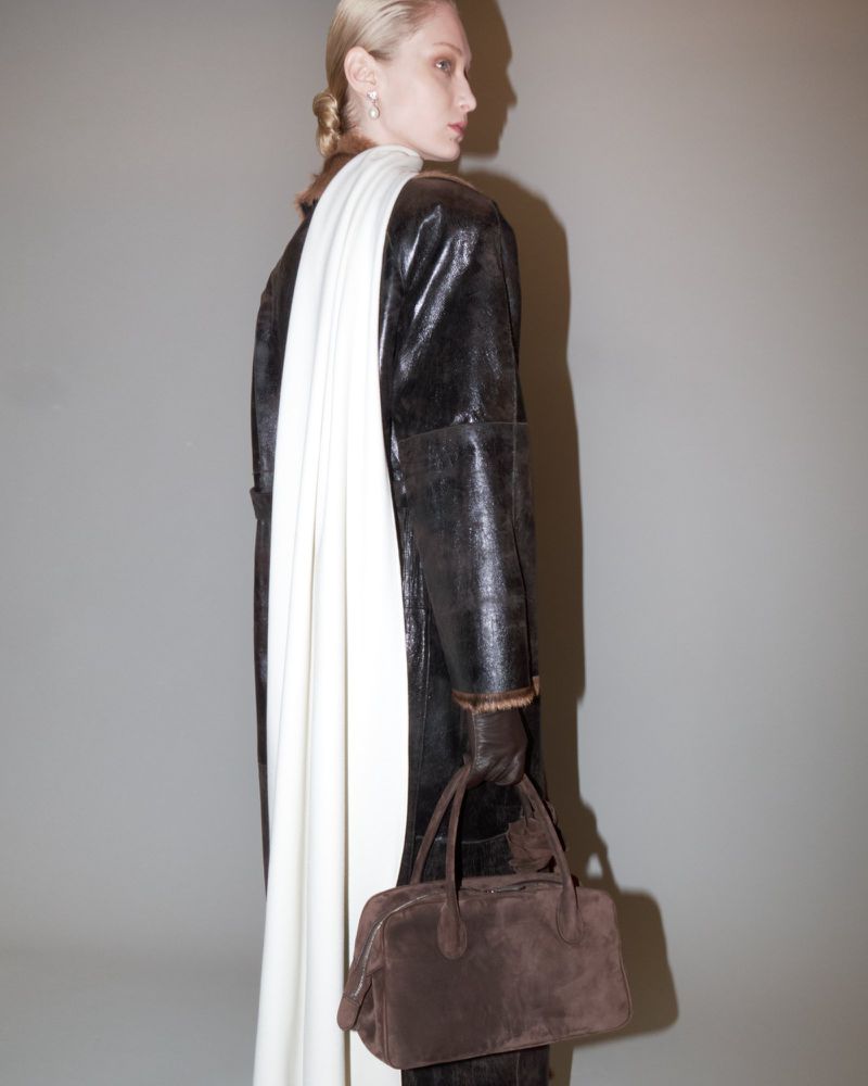 An adult woman in a dark overcoat poses in a studio with a cream-colored scarf and brown handbag, looking over her shoulder.