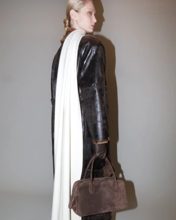 An adult woman in a dark overcoat poses in a studio with a cream-colored scarf and brown handbag, looking over her shoulder.