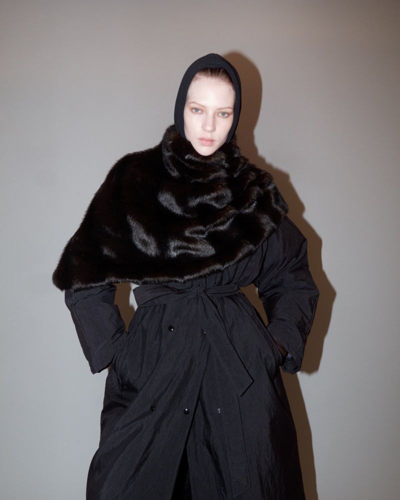 Woman wearing a black, hooded coat with a large, dark brown fur collar poses against a grey background in a studio setting.
