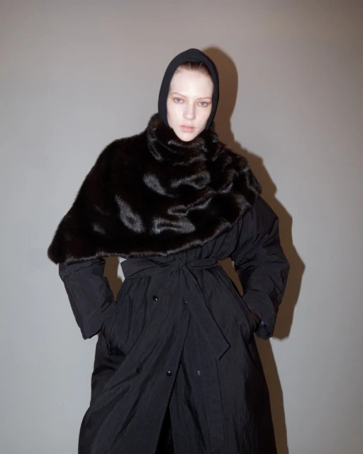 Woman wearing a black, hooded coat with a large, dark brown fur collar poses against a grey background in a studio setting.