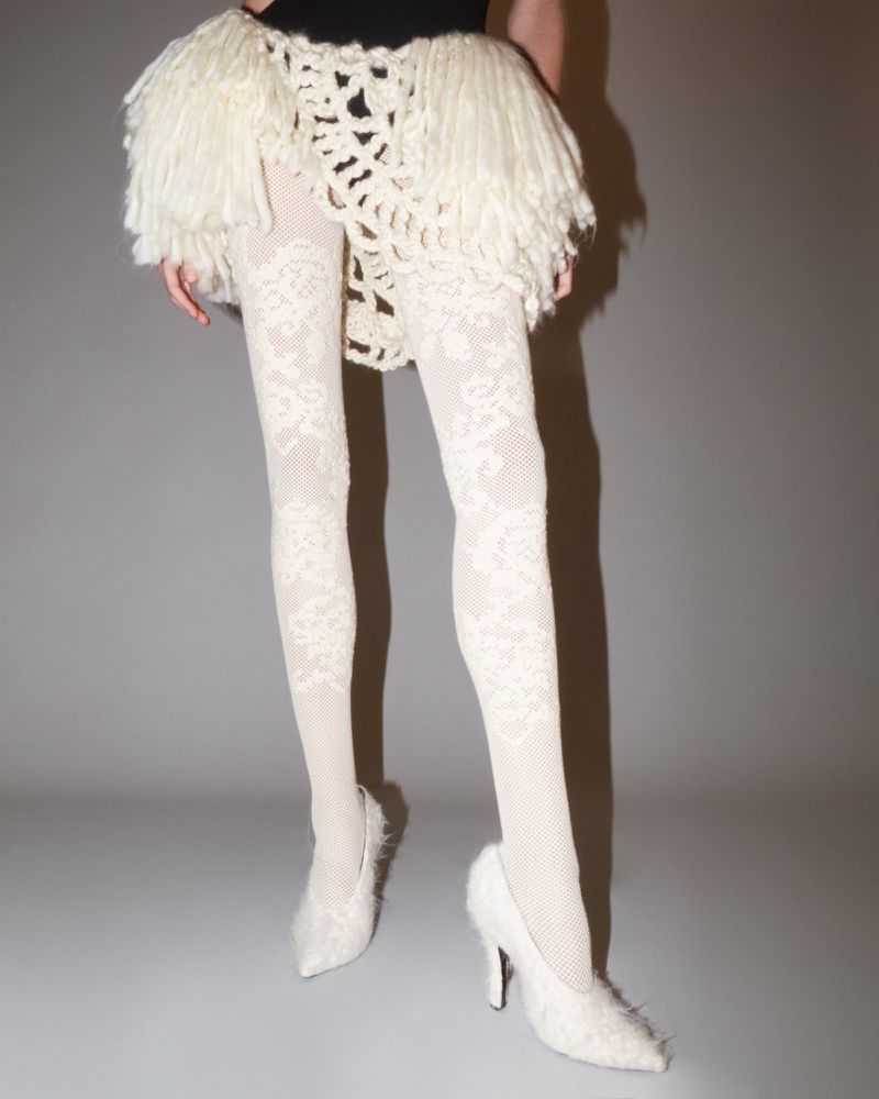 Fashionable woman wearing a black top with a fringed white skirt, patterned tights, and furry heeled shoes poses against a gray background.
