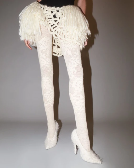 Fashionable woman wearing a black top with a fringed white skirt, patterned tights, and furry heeled shoes poses against a gray background.