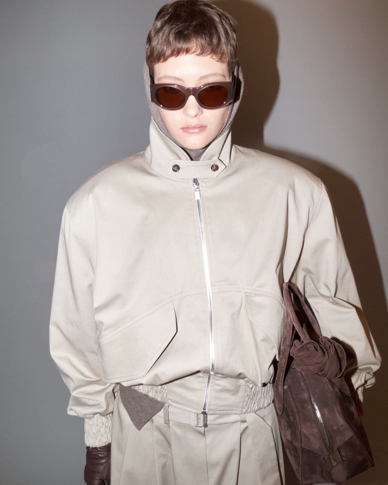 Woman in beige jacket, trousers, and sunglasses holds a brown leather purse against a grey background in a studio fashion shot.