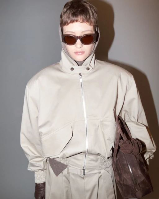 Woman in beige jacket, trousers, and sunglasses holds a brown leather purse against a grey background in a studio fashion shot.