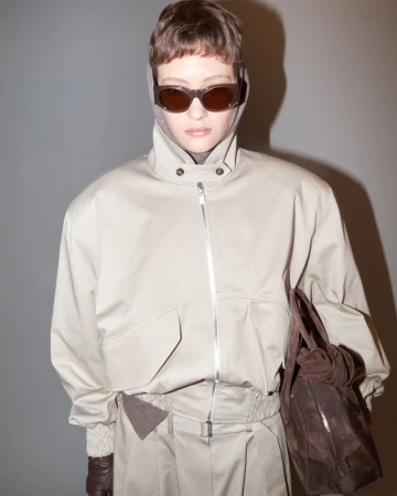 Woman in beige jacket, trousers, and sunglasses holds a brown leather purse against a grey background in a studio fashion shot.