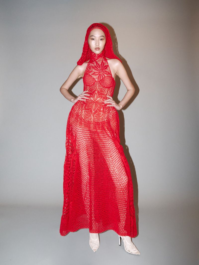 Woman in red crochet dress and matching head covering poses in a studio with hands on hips, showcasing high-fashion and style.