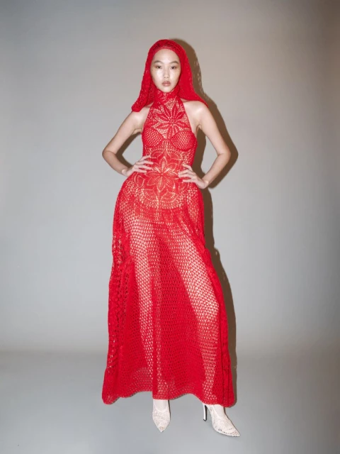 Woman in red crochet dress and matching head covering poses in a studio with hands on hips, showcasing high-fashion and style.