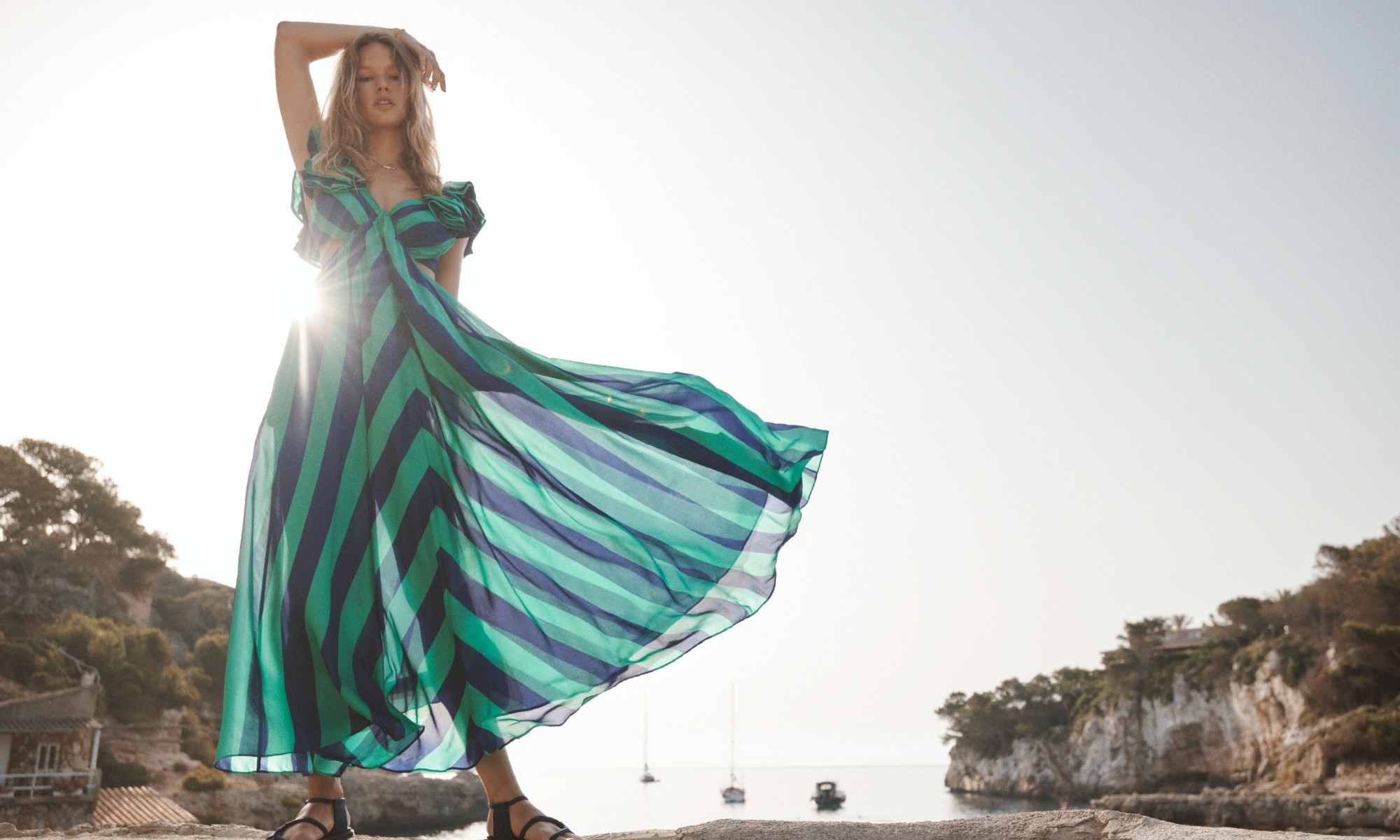 A woman in a flowing, striped dress poses on a rocky outcrop overlooking the ocean under a bright sky on a sunny day outdoors.