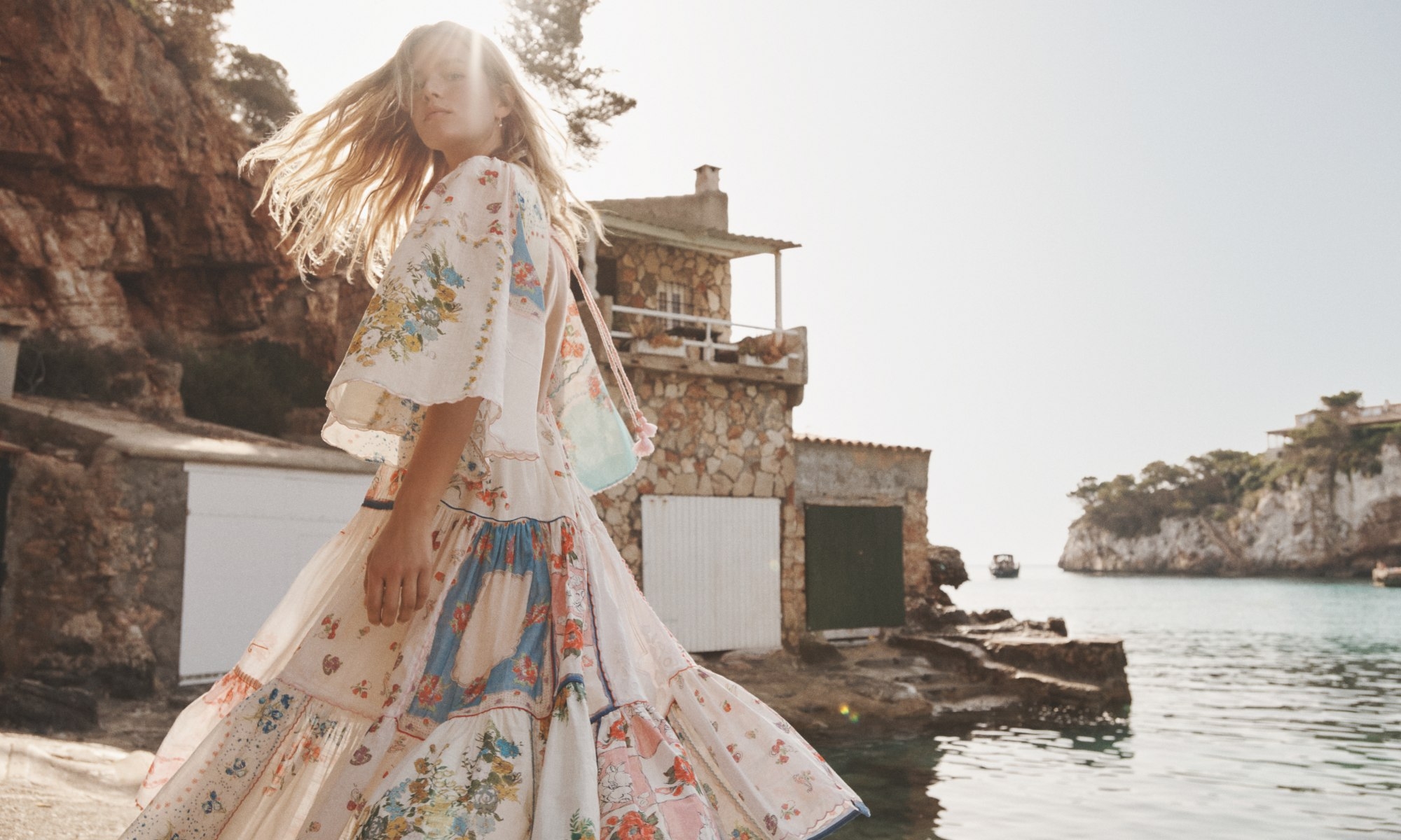 Woman in a floral dress stands on a rocky edge near the ocean with stone buildings and cliffs in the background on a sunny day.