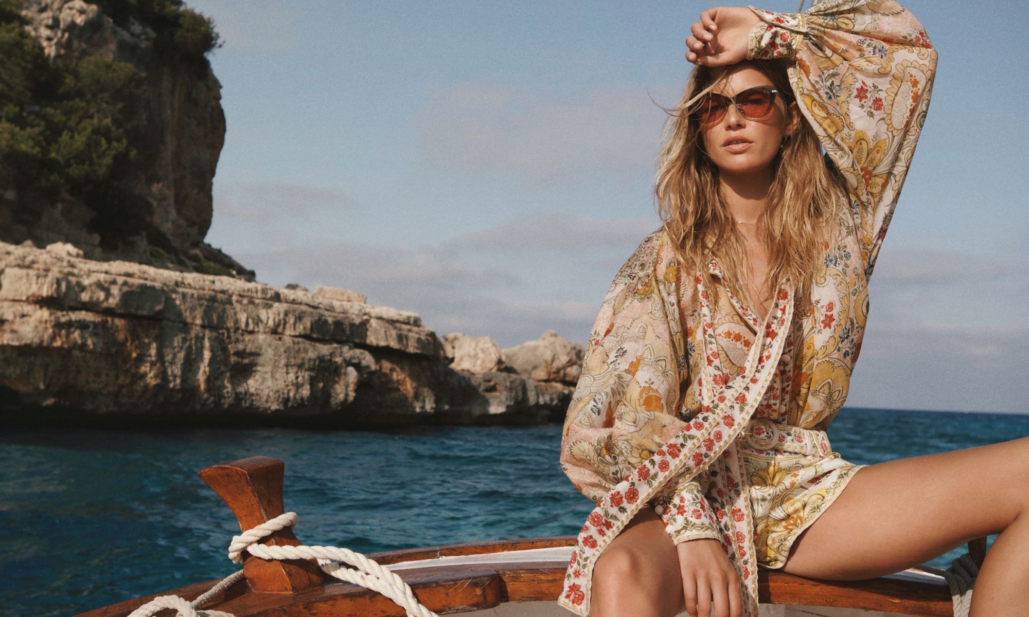 A blonde woman in a floral print outfit poses on a boat with a blue sea and rocky cliffs in the background on a sunny day.