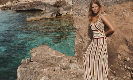 Model Anna Ewers in a striped knit dress leaning against rocky coast by ocean water for Zimmermann Resort Swim