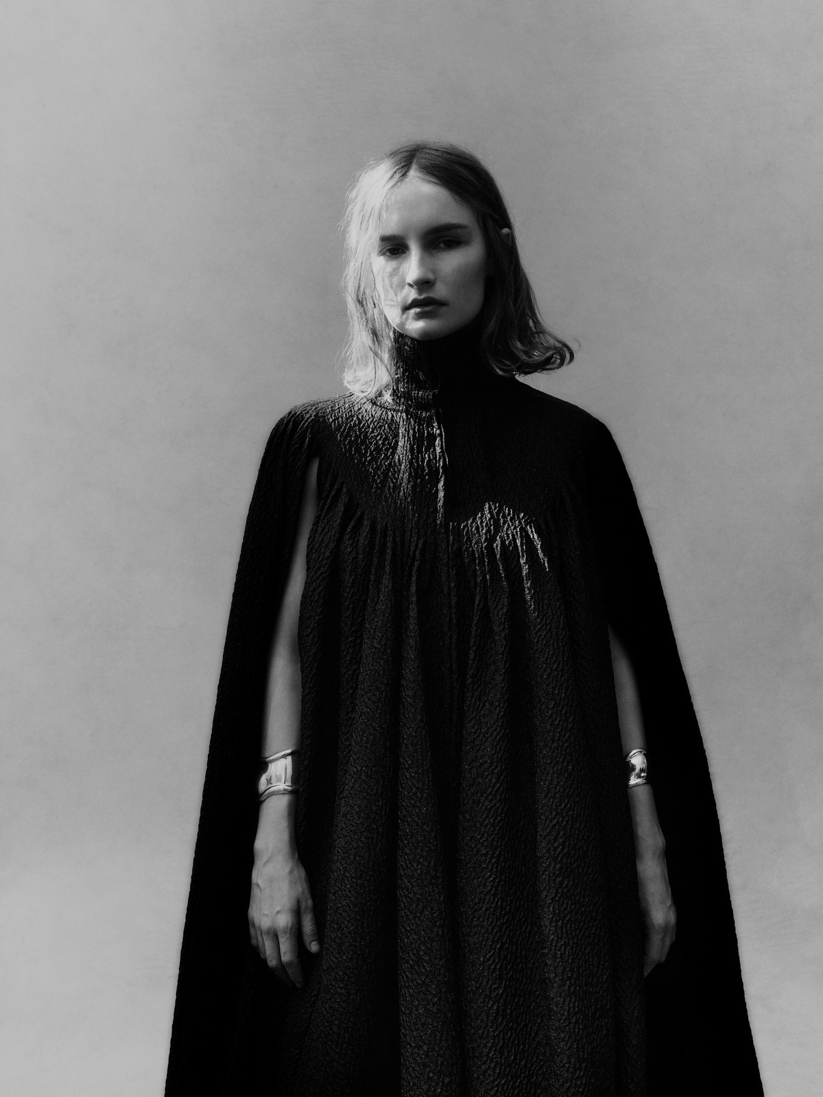 Monochrome portrait of a woman in a dark cape dress, posing in front of a soft grey background with natural light.