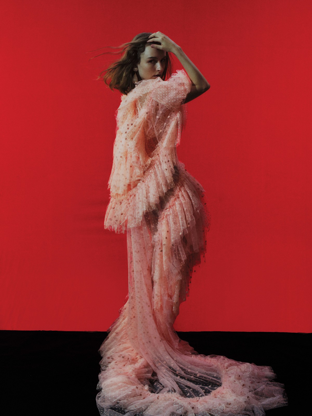 Fashion shot of a female model in a ruffled, light pink gown against a red backdrop and black floor, posing with her hand on her head.
