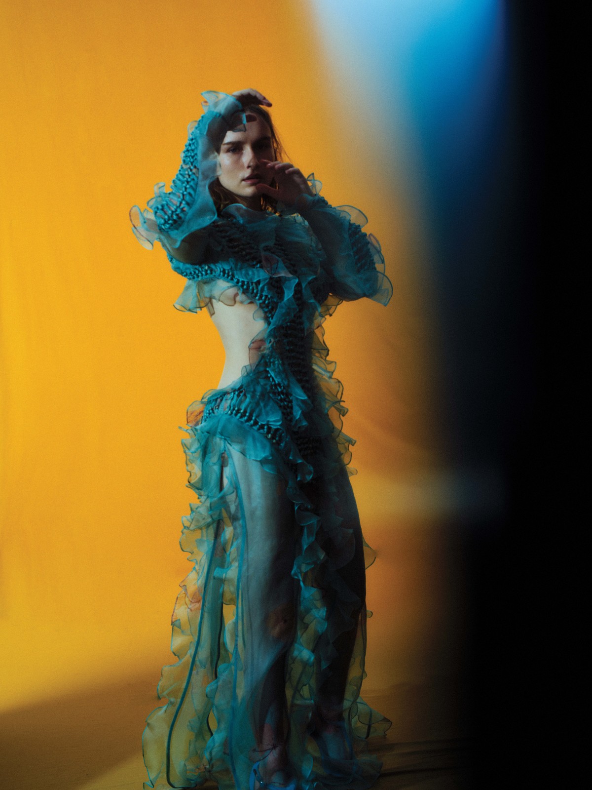 Woman in teal ruffled gown stands against yellow-to-black gradient background. Fashion, elegance, fabric, studio lighting, posing.