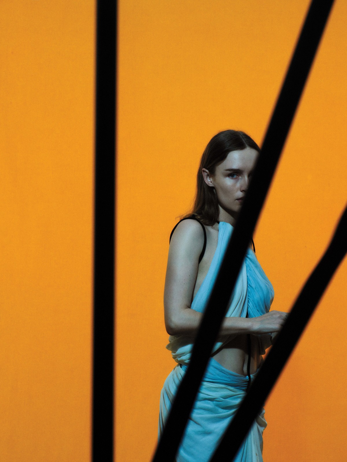 Woman with long hair poses in a draped blue garment against an orange backdrop with abstract black graphic elements, indoors.