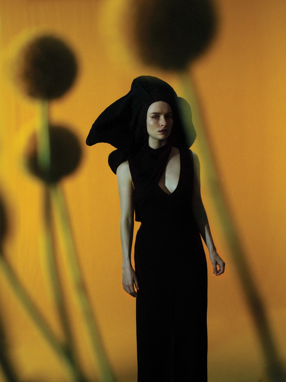 Woman poses in a black dress and flower-like hood, standing against a warm yellow background with blurred floral patterns.