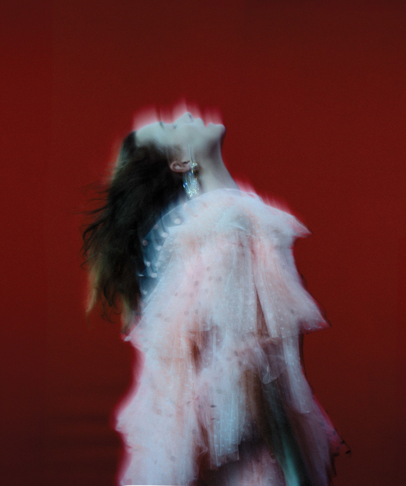 Woman with blurred motion posing in layered tulle dress against a vivid red backdrop, featuring jewelry, for beauty or fashion editorial.