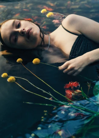 Woman in dark water with wildflowers, face up, wearing black top and necklace, portrait-style. Ethereal and moody.