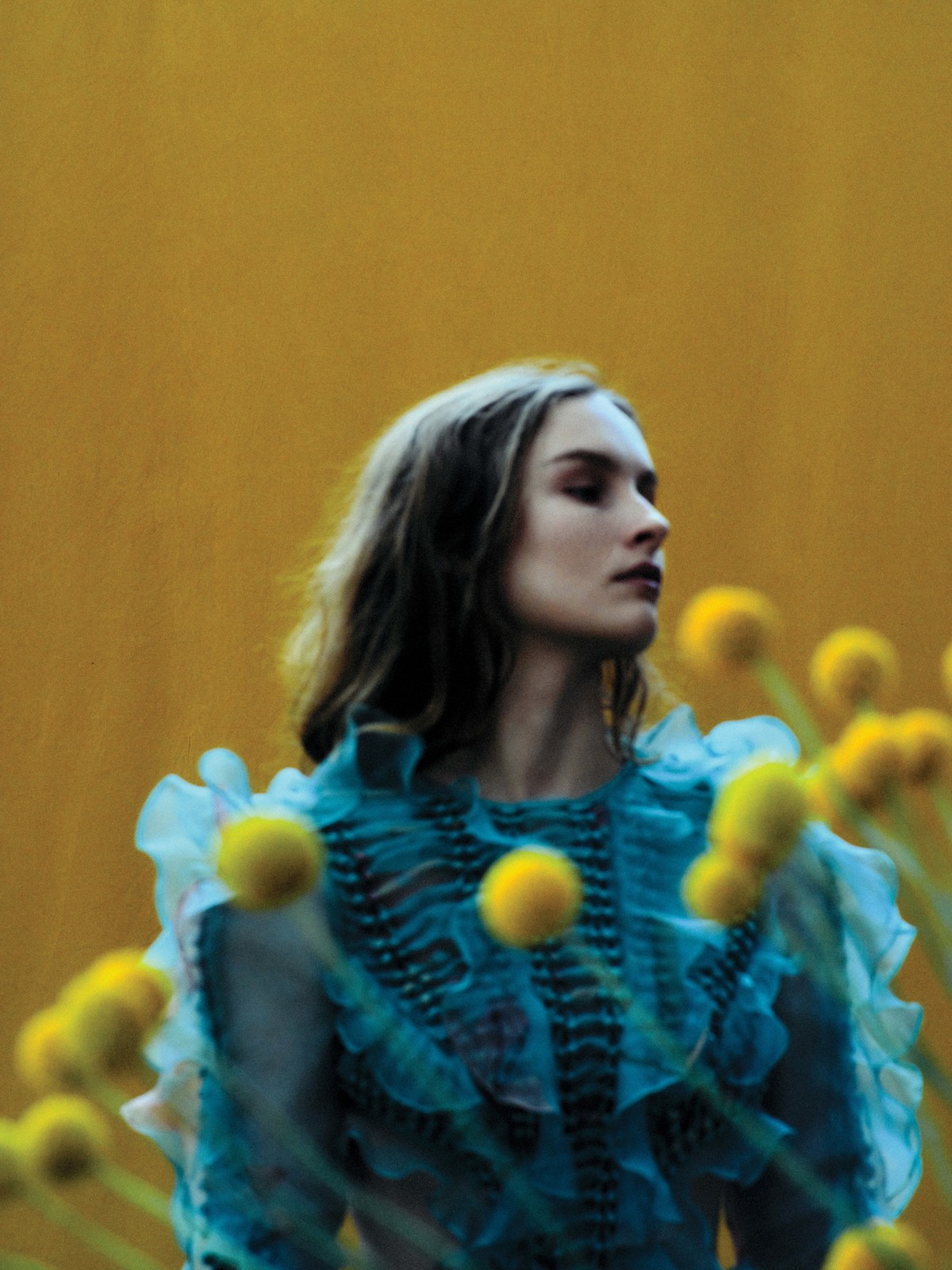 Portrait of a woman in a teal dress with ruffle details, posed among yellow flowers against a solid yellow backdrop in an outdoor setting.