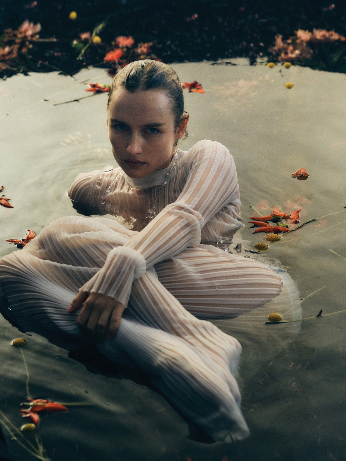 Olivia De Jonge wearing a white sheer pleated top seated in water with floating flower petals for T Magazine
