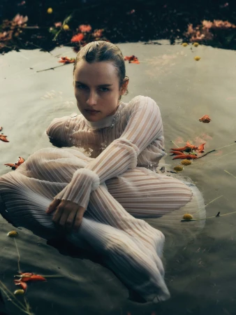 Olivia De Jonge wearing a white sheer pleated top seated in water with floating flower petals for T Magazine