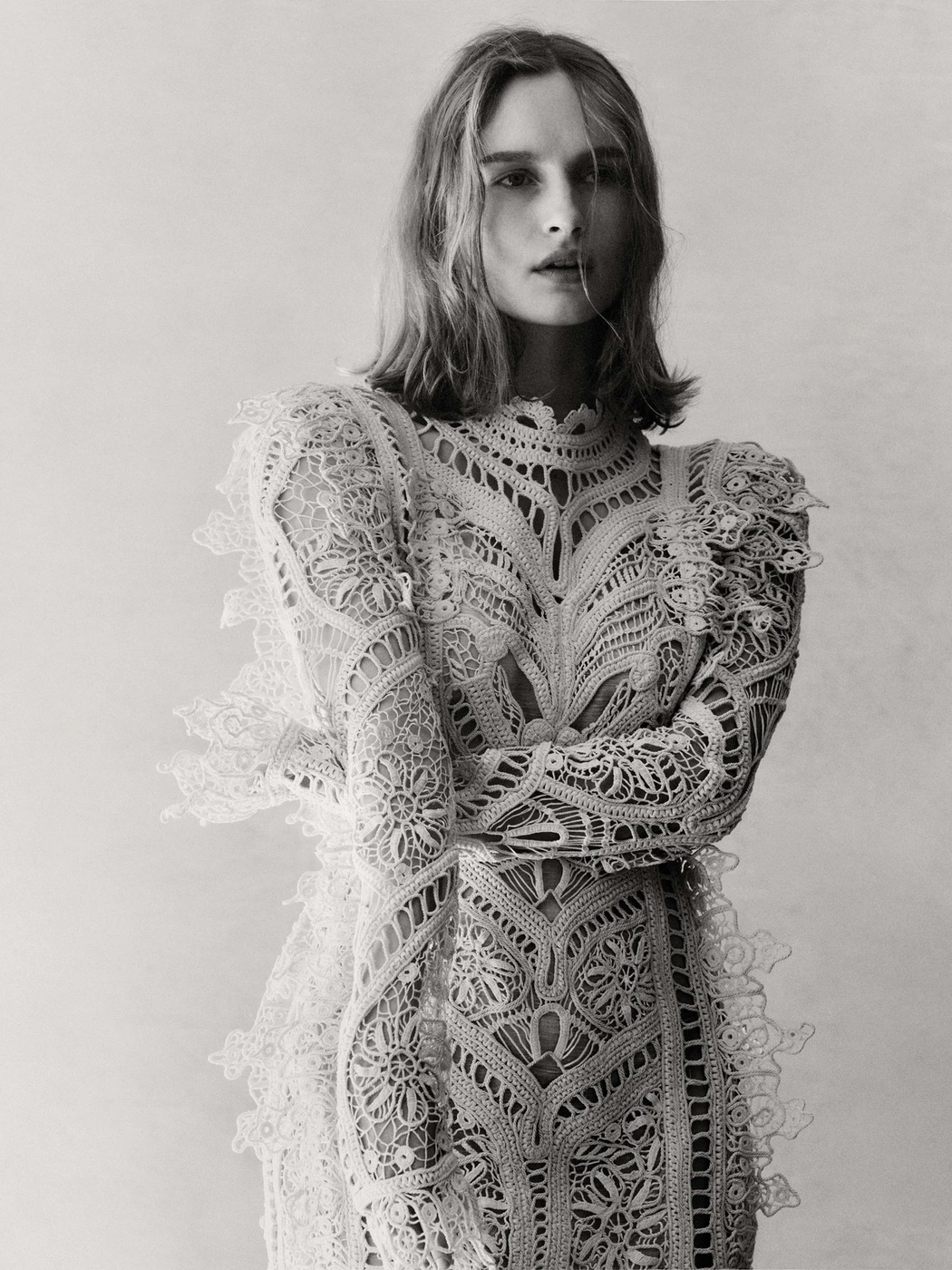 Olivia De Jonge wearing an intricate white lace long sleeve dress against a plain studio background in a portrait photograph by Georges Antoni.