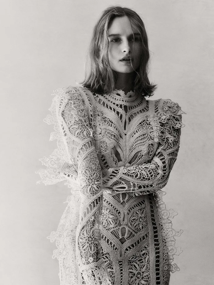 Olivia De Jonge wearing an intricate white lace long sleeve dress against a plain studio background in a portrait photograph by Georges Antoni.