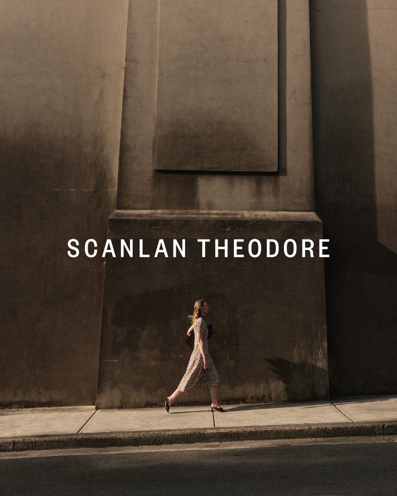 Woman in dress walking past weathered concrete wall with 'SCANLAN THEODORE' text overlay; urban style, fashion editorial.