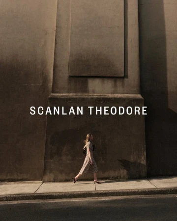 Woman in dress walking past weathered concrete wall with 'SCANLAN THEODORE' text overlay; urban style, fashion editorial.