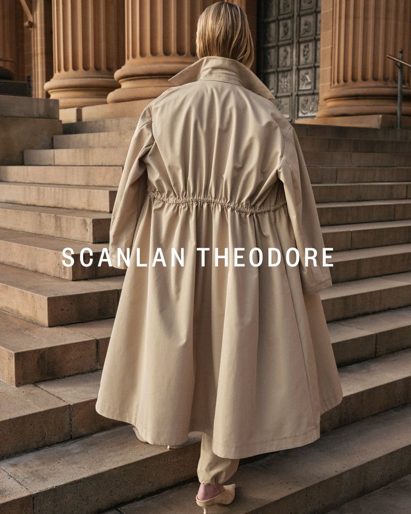 Woman in a tan trench coat walks up stone steps framed by large columns, showcasing fashion and architecture in an outdoor setting.