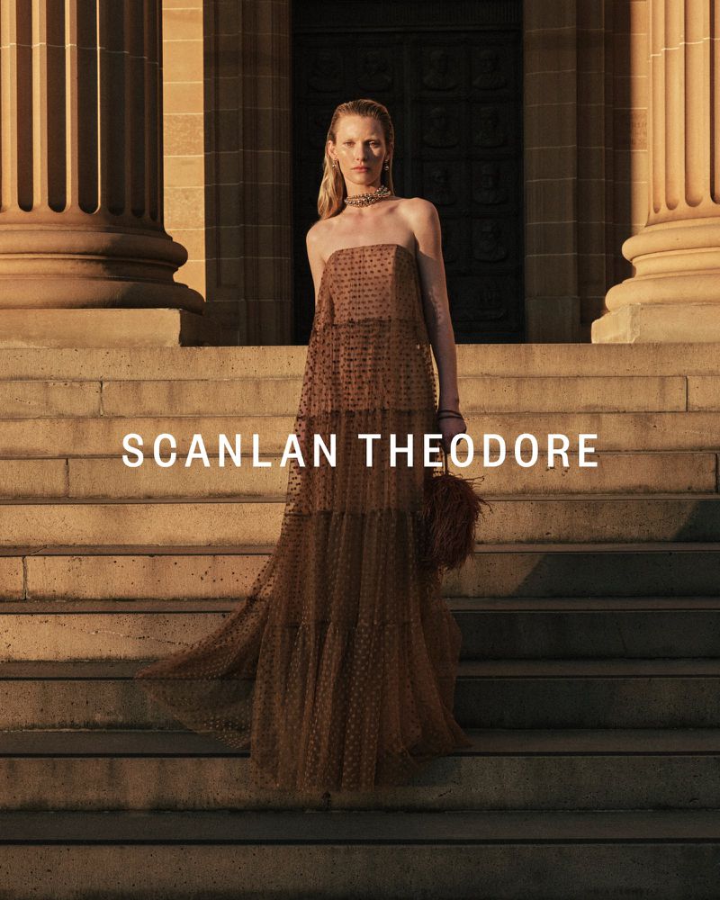 A blonde woman in a brown dress poses on stone steps in front of a building with pillars, wearing a necklace and holding a feathered bag.