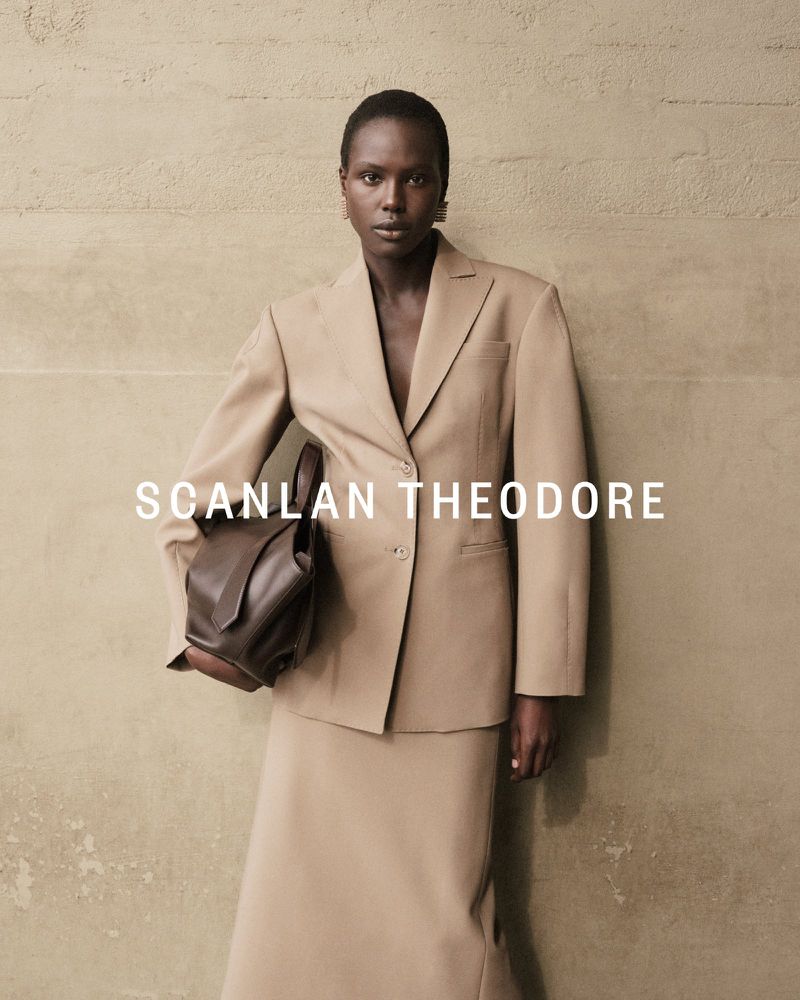 A stylish woman in a beige suit holds a dark brown leather bag against a textured wall, showcasing fashion and elegance.