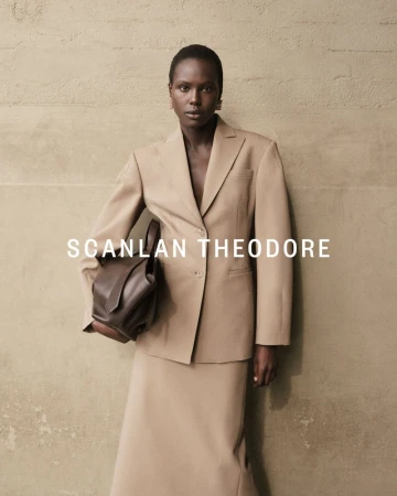 A stylish woman in a beige suit holds a dark brown leather bag against a textured wall, showcasing fashion and elegance.