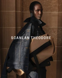 Model wearing a dark denim jacket and holding a tan leather bag against an architectural stone wall in campaign imagery.