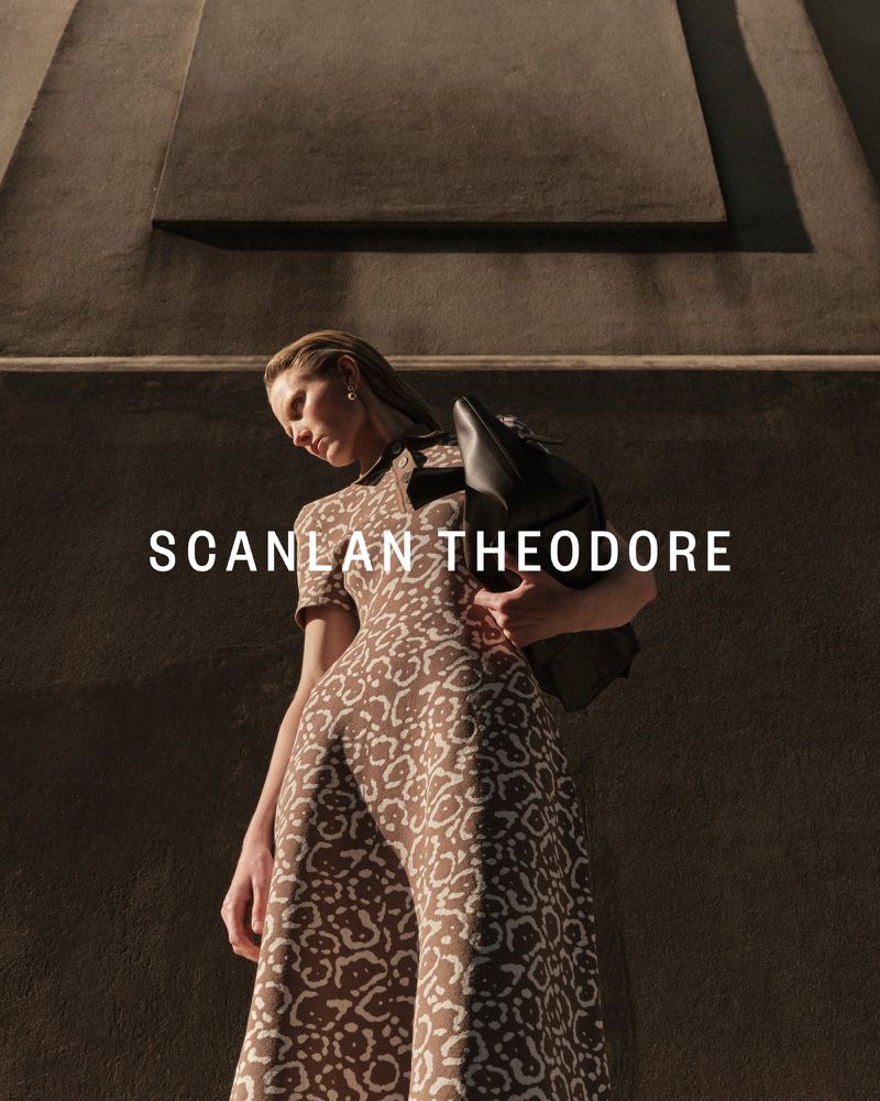Model in patterned dress holding a black handbag standing against a textured dark architectural background for Scanlan Theodore